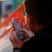 A person holding and interacting with a mobile device, with a focus on the screen displaying colorful graphics. The background contains a blurred view of an outdoor setting with a grid-like orange structure.
