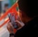 A person holding and interacting with a mobile device, with a focus on the screen displaying colorful graphics. The background contains a blurred view of an outdoor setting with a grid-like orange structure.